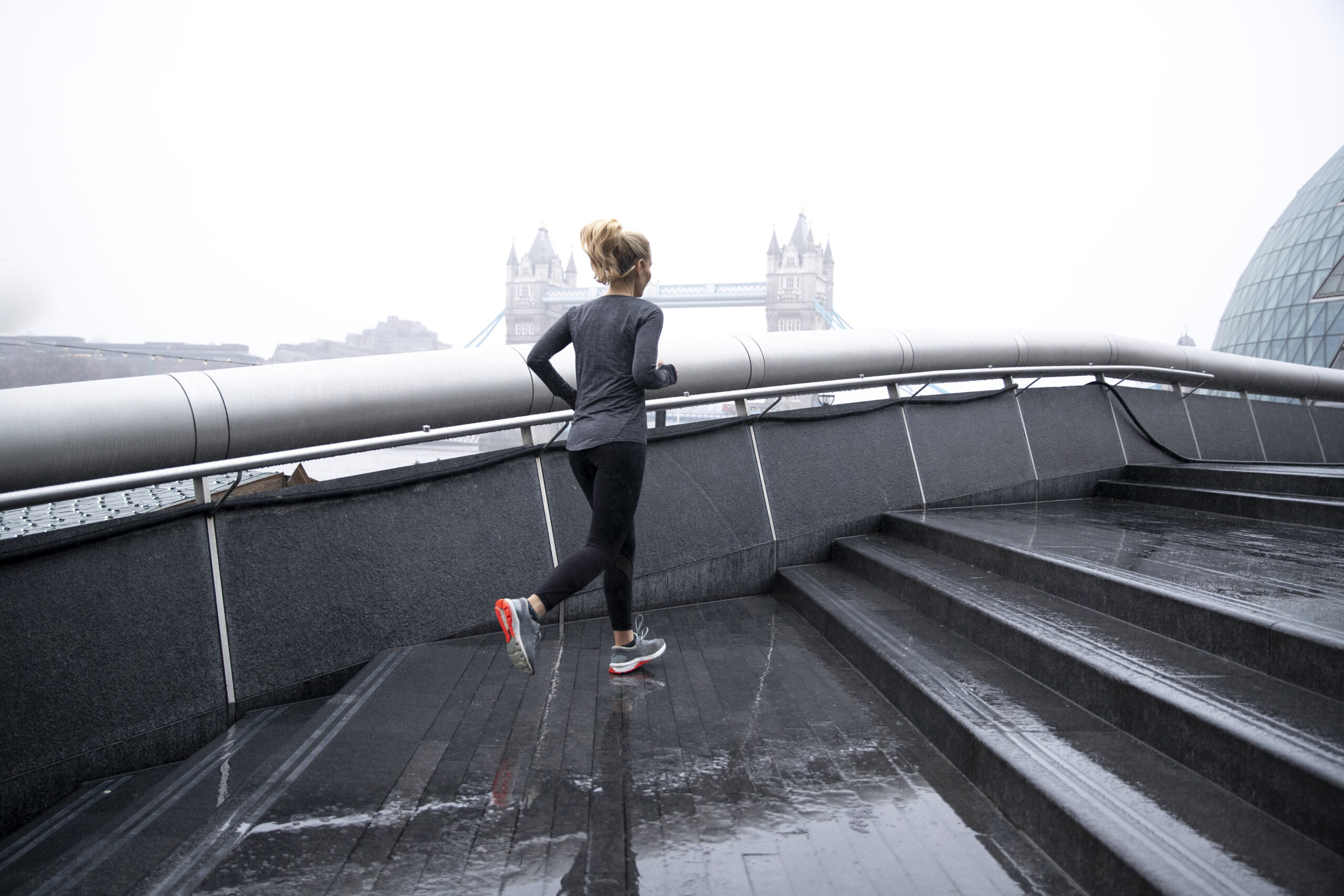 Woman running city streets up stairs while it rains. Bridge in background
